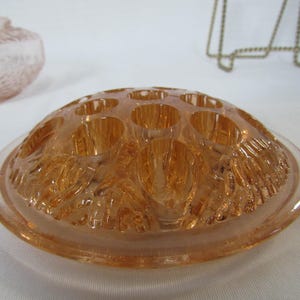 May include: A vintage, peach-colored glass flower frog with multiple holes for arranging stems. The frog has a rounded, dome-like shape with a textured, floral design on the underside. The rim is smooth and clear.