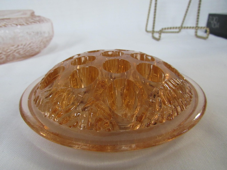May include: A vintage, peach-colored glass flower frog with multiple holes for arranging stems. The frog has a rounded, dome-like shape with a textured, floral design on the underside. The rim is smooth and clear.