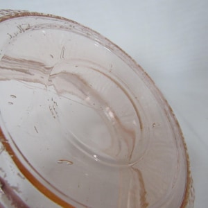 May include: A clear, light pink glass object with a textured rim and a circular base. The glass has visible imperfections and bubbles, with a slightly amber hue around the edges. The object appears to be a vintage piece.
