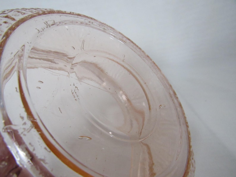 May include: A clear, light pink glass object with a textured rim and a circular base. The glass has visible imperfections and bubbles, with a slightly amber hue around the edges. The object appears to be a vintage piece.