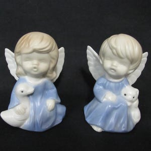 May include: Two porcelain angel figurines with wings, both wearing blue dresses. One angel holds a white goose, the other holds a white cat.