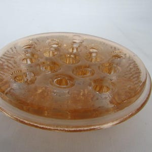May include: A vintage, peach-colored glass flower frog. The round, shallow dish has multiple holes for arranging flower stems. The rim is slightly raised and the glass has a textured pattern. A classic floral arranging tool.