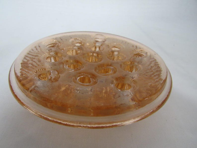 May include: A vintage, peach-colored glass flower frog. The round, shallow dish has multiple holes for arranging flower stems. The rim is slightly raised and the glass has a textured pattern. A classic floral arranging tool.