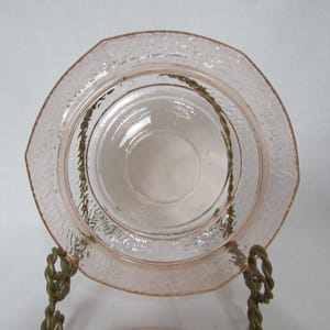 May include: A set of pink glass plates, one with an octagonal shape and textured rim, and a smaller, round plate nested inside. The plates are displayed on a decorative brass stand, showcasing vintage tableware.