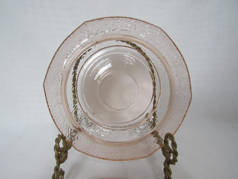 May include: A set of pink glass plates, one with an octagonal shape and textured rim, and a smaller, round plate nested inside. The plates are displayed on a decorative brass stand, showcasing vintage tableware.