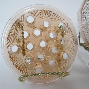 May include: A clear, round, peach-colored glass flower frog with multiple holes for arranging stems. The frog has a decorative pattern and is displayed on a brass wire stand. A second glass object is partially visible.