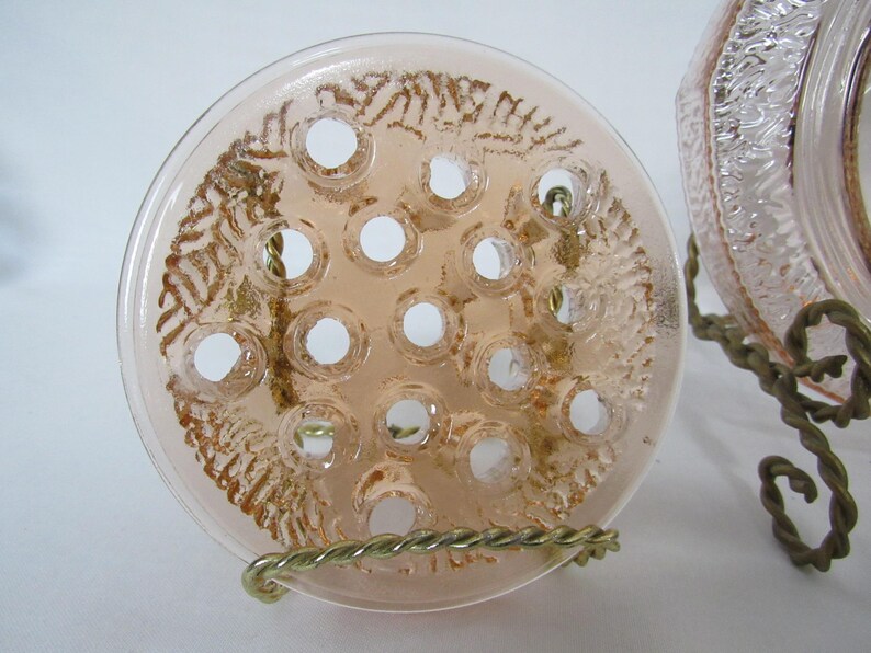 May include: A clear, round, peach-colored glass flower frog with multiple holes for arranging stems. The frog has a decorative pattern and is displayed on a brass wire stand. A second glass object is partially visible.