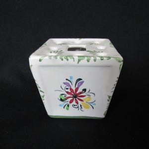 May include: White ceramic toothbrush holder with a floral design. The holder has a square shape with a green border and a colorful flower in the center. The top has several holes for toothbrushes.
