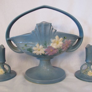 May include: A blue ceramic set of a basket with a handle and two matching candle holders. The basket and candle holders are decorated with white and pink flowers.
