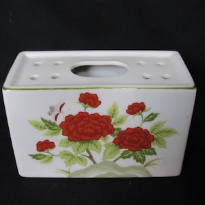 May include: White ceramic flower vase with a floral design featuring red flowers and green leaves. The vase has a rectangular shape with a small oval opening on the top and multiple holes on the sides.