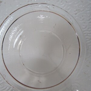 May include: Clear glass plate with a raised, circular center and a decorative, textured rim. The rim features a subtle, brown-toned accent. The plate's design is simple and elegant, suitable for serving or display.