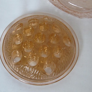 May include: A vintage, peach-colored glass flower frog with multiple holes for arranging stems. The frog has a textured, circular design and is likely used for floral arrangements. Another piece of peach-colored glass is visible in the background.