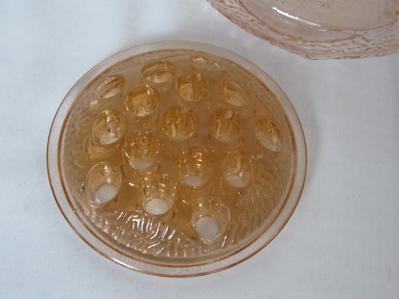 May include: A vintage, peach-colored glass flower frog with multiple holes for arranging stems. The frog has a textured, circular design and is likely used for floral arrangements. Another piece of peach-colored glass is visible in the background.