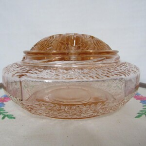 May include: A vintage, peach-colored glass jar with a textured exterior and a domed lid. The jar has a round base and a textured pattern around the sides. The lid features a floral design. The jar is likely used for storage or display.