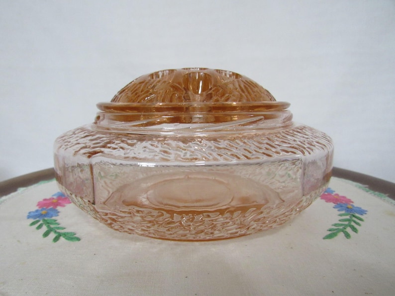 May include: A vintage, peach-colored glass jar with a textured exterior and a domed lid. The jar has a round base and a textured pattern around the sides. The lid features a floral design. The jar is likely used for storage or display.