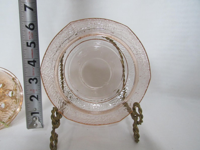 May include: A vintage pink glass saucer with a textured rim and a central depression, displayed on a decorative brass stand. The saucer is approximately 4 inches in diameter. The glass has a delicate, translucent quality.