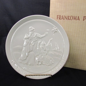 May include: White ceramic plate with a raised design of three figures, a woman and two children, with a star above them. The plate has the text "Is the Word" on the left and "Rejoice" on the right.