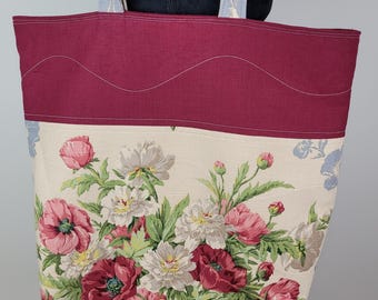 May include: A large tote bag with a floral pattern featuring pink and white flowers on an off-white background. The top of the bag is a solid burgundy color with a wavy stitched design. The handles are a light blue and white floral pattern.