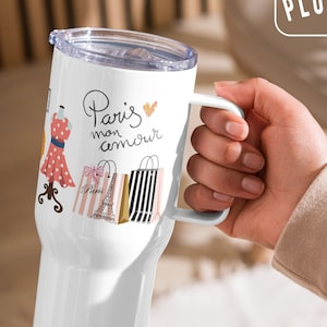May include: White travel mug with a handle. The mug has a design of a dress, Eiffel Tower, and shopping bags with the text "Paris mon amour" and a pink heart.