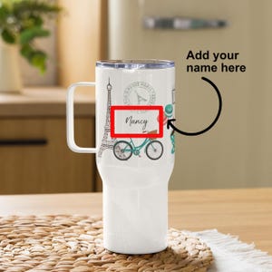 May include: White travel mug with a design featuring the Eiffel Tower, a bicycle, and the words "Paris Paris Paris". The mug has a black handle and a red box with the word "Nancy" in it.