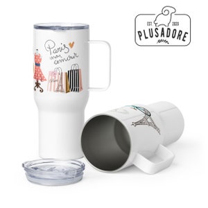 May include: Two white travel mugs with a black handle. One mug features a graphic of a dress, shopping bags, and the text "Paris mon amour". The other mug features a graphic of the Eiffel Tower. Both mugs have a clear lid.