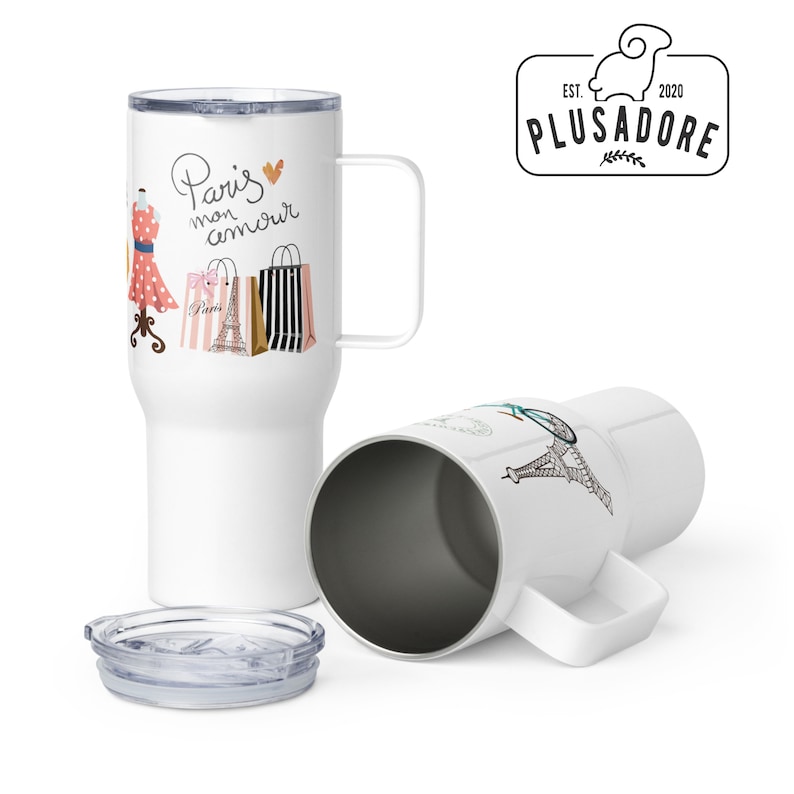 May include: Two white travel mugs with a black handle. One mug features a graphic of a dress, shopping bags, and the text "Paris mon amour". The other mug features a graphic of the Eiffel Tower. Both mugs have a clear lid.