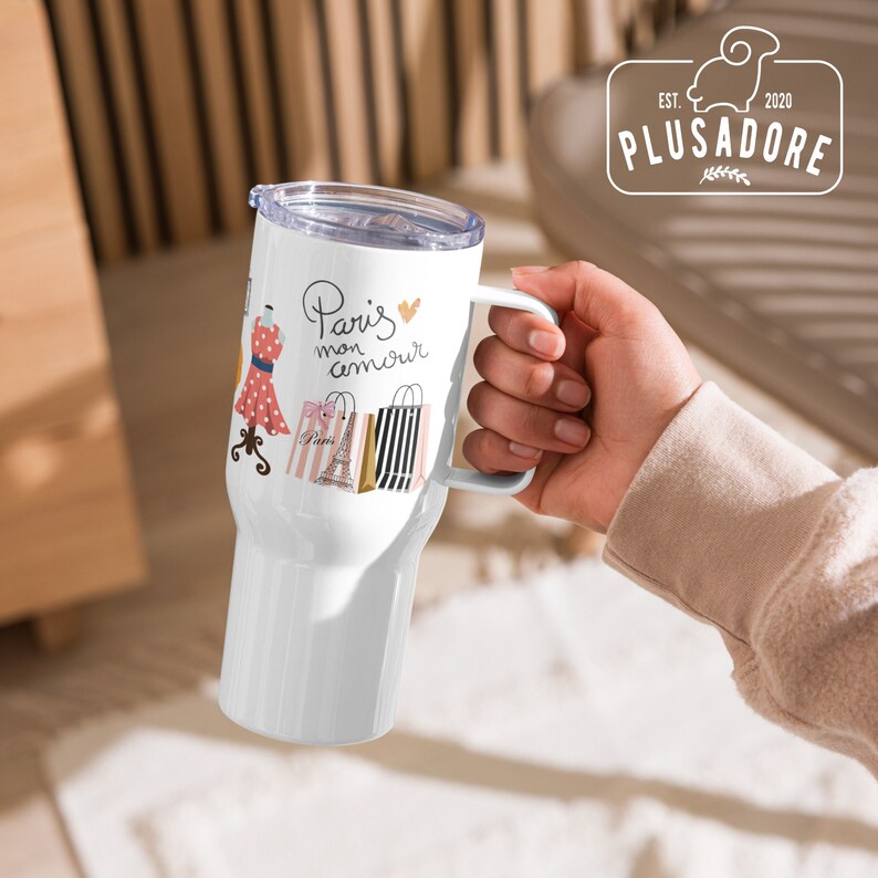 May include: White travel mug with a handle. The mug has a design of a dress, Eiffel Tower, and shopping bags with the text "Paris mon amour" and a pink heart.