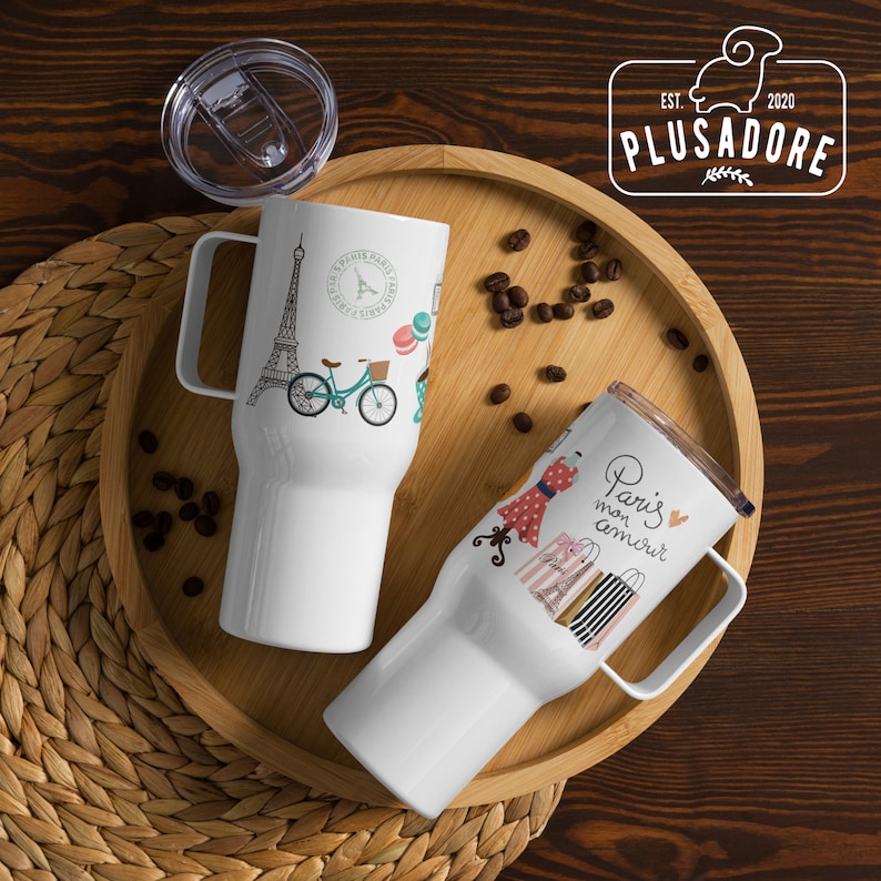 May include: Two white travel mugs with different designs. The first mug features a black and white illustration of the Eiffel Tower, a bicycle, and the words "Paris Paris Paris". The second mug features a red dress, shopping bags, and the words "Paris mon amour".