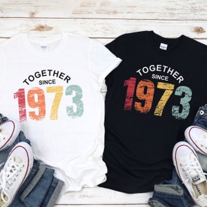 May include: Two t-shirts with a vintage-style graphic that says "Together Since 1973". The t-shirts are white and black, and the graphic is in rainbow colors with a distressed look.