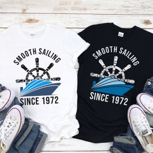 May include: Two t-shirts with a graphic design of a boat with a steering wheel and the text "Smooth Sailing Since 1972". One t-shirt is white and the other is black.