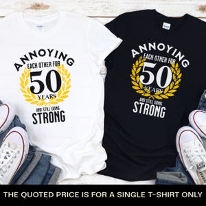 May include: Two t-shirts with a black and white design. The text on the shirts reads "Annoying Each Other For 50 Years And Still Going Strong". The shirts are laid out on a wooden surface with jeans and sneakers.