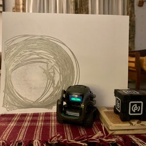 May include: A black and green robot with a light on its face, a white piece of paper with a gray pencil drawing of a circle, and a black cube with the letter "P" on it.