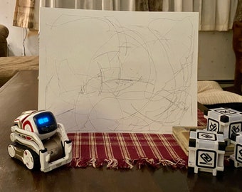 May include: A white and red robot with gold accents is on a table in front of a white board with scribbles on it. Three white cubes with black accents are on the table.