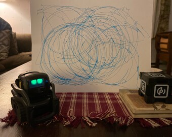 May include: A black and gray robot with green lights on its face is sitting on a table with a red and white plaid tablecloth. A small black cube with a white circle and a white letter 'G' is sitting on a stack of books.