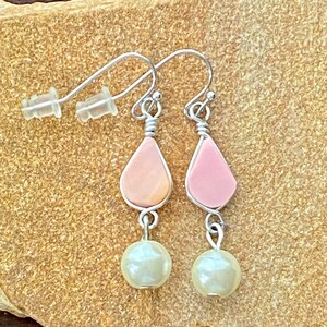 May include: Dangle earrings featuring a teardrop-shaped pink and silver design, with a silver wire wrap. Each earring has a cream-coloured pearl bead hanging below. The earrings are displayed on a textured, tan surface.