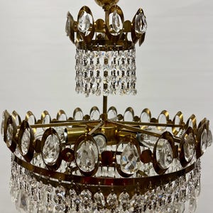 Op de afbeelding: Een messing en kristallen kroonluchter met een cirkelvormig ontwerp en meerdere lagen kristallen druppels. De kroonluchter heeft een gouden afwerking en beschikt over een centrale koepel met een decoratief ontwerp.