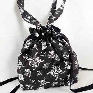 Puede incluir: Bolsa de cordón negra con un patrón de calaveras y huesos cruzados blancos. La bolsa tiene dos correas largas negras.