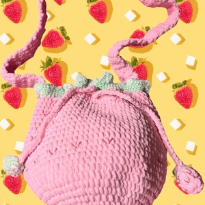 Puede incluir: Un bolso de ganchillo rosa de forma redonda con cierre de cord&oacute;n y una superficie texturizada. El bolso tiene un borde con volantes de color verde claro en la abertura superior y una correa larga de ganchillo rosa a juego. Dos peque&ntilde;os pompones de ganchillo verde claro cuelgan de los cordones a ambos lados del bolso.