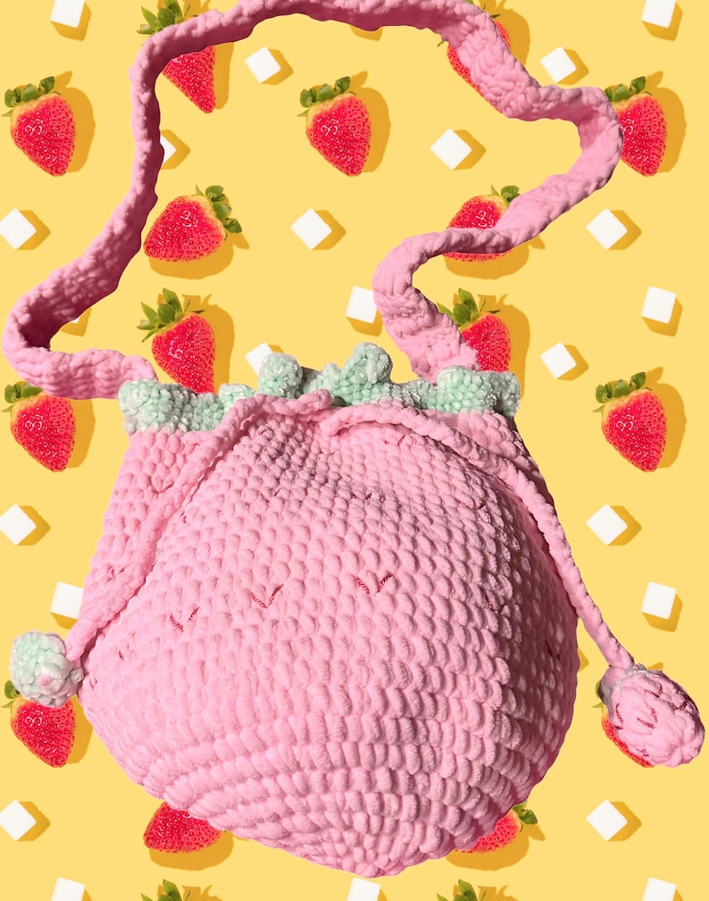 Puede incluir: Un bolso de ganchillo rosa de forma redonda con cierre de cord&oacute;n y una superficie texturizada. El bolso tiene un borde con volantes de color verde claro en la abertura superior y una correa larga de ganchillo rosa a juego. Dos peque&ntilde;os pompones de ganchillo verde claro cuelgan de los cordones a ambos lados del bolso.
