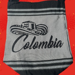 Puede incluir: Bolso de hombro negro y gris con la palabra "Colombia" en cursiva. El bolso presenta un gráfico de sombrero y rayas horizontales. La correa es negra.
