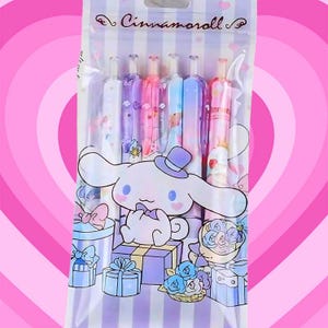 Puede incluir: Un juego de seis bol&iacute;grafos de gel en una bolsa de pl&aacute;stico transparente con la marca Cinnamoroll. El empaque muestra un personaje de perro blanco, cajas de regalo e ilustraciones florales en tonos pastel sobre un fondo de rayas.