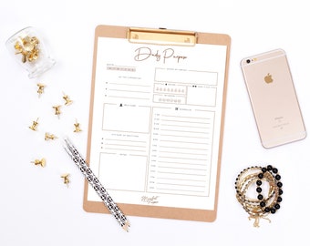 May include: A printable daily planner on a clipboard with a brown background. The planner includes sections for daily goals, self-care, gratitude, and a schedule. The planner is surrounded by office supplies, including gold push pins, a pencil, and a smartphone.