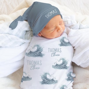 May include: A newborn baby wrapped in a white swaddle blanket with a wave pattern and the name "Thomas Ethan" printed on it. The baby wears a matching blue hat with the name "Thomas" on it. The baby is lying on a fluffy white blanket.