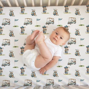 May include: A baby in a white onesie lies on a crib sheet with a golf theme. The sheet features a repeating pattern of cartoon golfers, golf carts, golf bags, and flags in shades of blue, green, and tan on a white background.