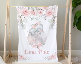 May include: A white baby blanket featuring a watercolor illustration of a gray elephant sleeping on a gray sphere, surrounded by pink roses and greenery. The name "Luna Mae" is written in pink script. The blanket is displayed on a wooden drying rack.