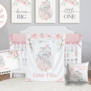 May include: A pink and gray baby crib bedding set with a floral design and a sleeping elephant. The set includes a crib sheet, a blanket, a pillow, and a pillowcase. The blanket and pillowcase feature the text "Luna Mae". The pillow features the text "Sweet Dreams Little One".