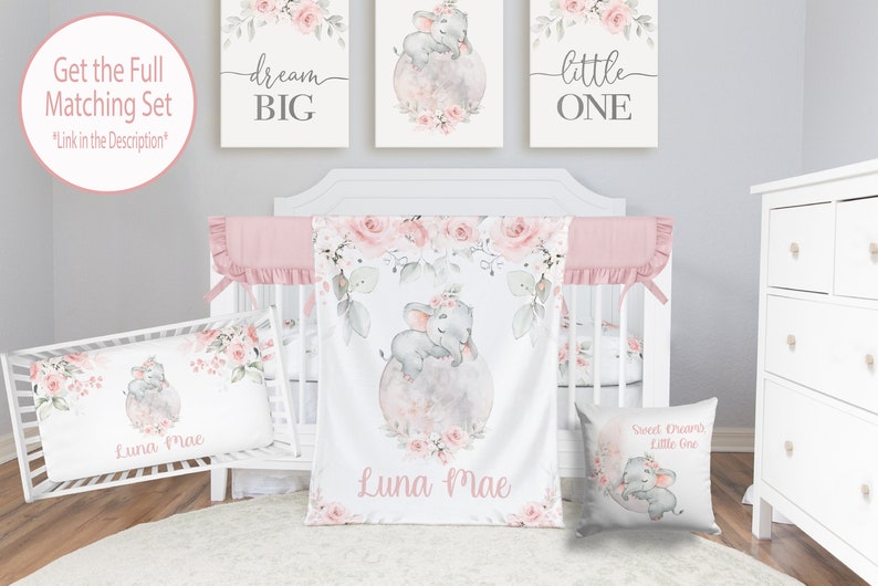 May include: A pink and gray baby crib bedding set with a floral design and a sleeping elephant. The set includes a crib sheet, a blanket, a pillow, and a pillowcase. The blanket and pillowcase feature the text "Luna Mae". The pillow features the text "Sweet Dreams Little One".