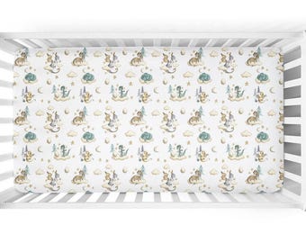 May include: White fitted crib sheet with a repeating pattern of watercolor dragons, clouds, stars, and moons in shades of brown, green, and yellow. The sheet is inside a white crib.