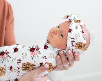 May include: A newborn baby wrapped in a white swaddle blanket and matching headband, both adorned with a floral pattern in burgundy and pink. The name "Madison" is printed on the fabric, along with a brown cow illustration.