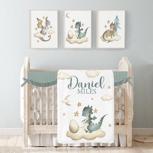 Puede incluir: Una escena de guardería con una cuna y tres impresiones enmarcadas en acuarela. La manta blanca tiene el nombre "Daniel Miles" y una ilustración de dragón. Las impresiones representan dragones, nubes y estrellas.